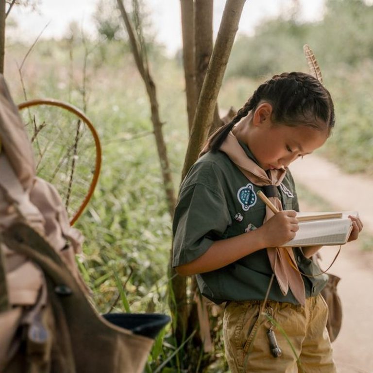 Asian girl scout writing in notebook outdoors, capturing nature notes.