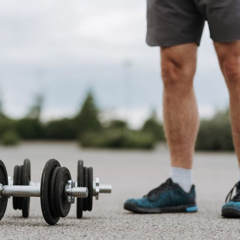 Crop anonymous male in sportswear standing near heavy metal dumbbells on asphalt road