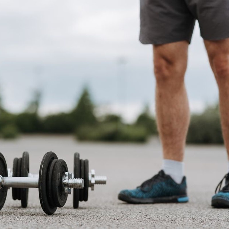 Crop anonymous male in sportswear standing near heavy metal dumbbells on asphalt road