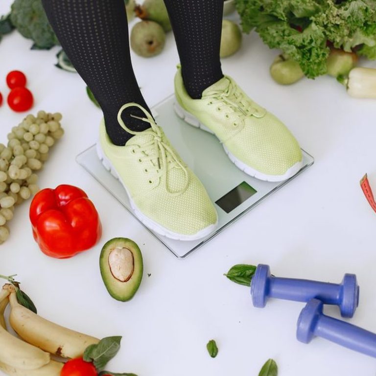 Person standing on scales surrounded by fresh produce and fitness equipment symbolizing a balanced lifestyle.