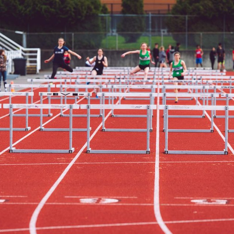 Women athletes compete in a hurdles race at a Victoria track event on a sunny day.