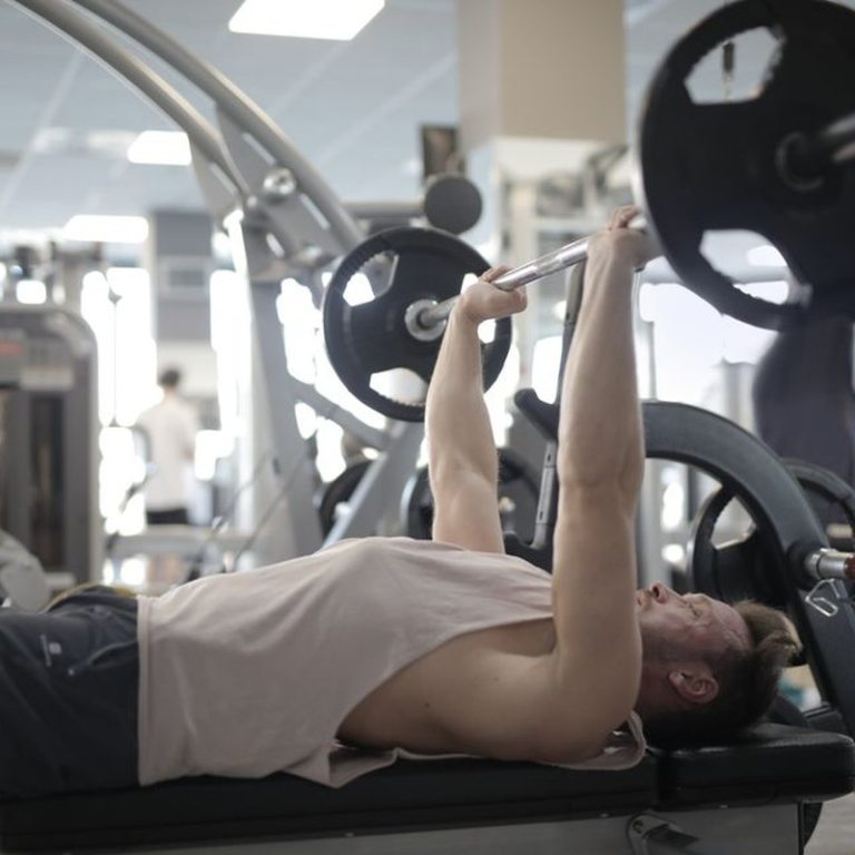 Side view of strong young sportsman in casual sportswear pressing heavy barbell on bench while intense training in modern fitness center