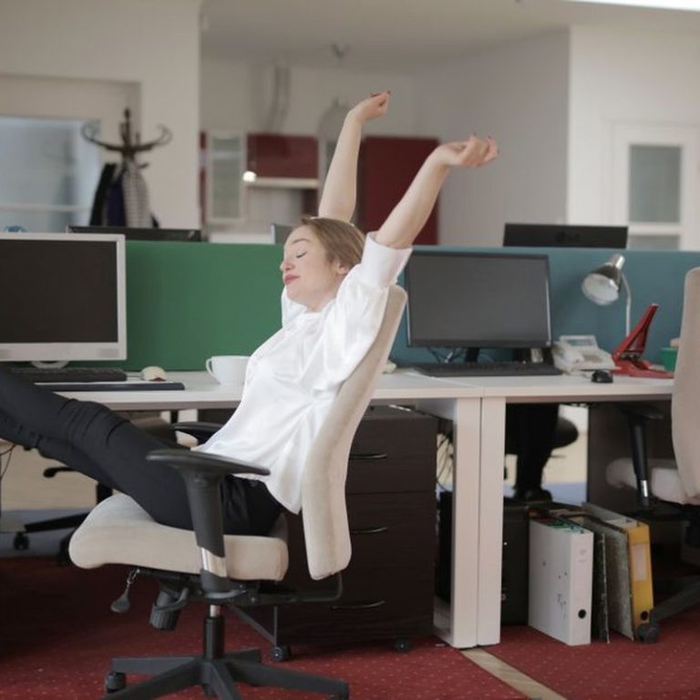Side view of cheerful female employee in formal outfit sitting on office chair with crossed legs on desk and stretching while resting during work with closed eyes