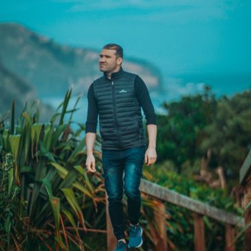A man walks along a scenic coastal path surrounded by lush greenery and a vibrant blue sky.
