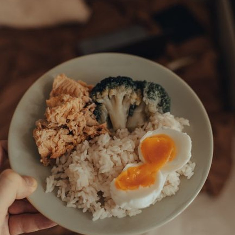 From above of crop anonymous person holding plate with homemade lunch including fried fish with broccoli and boiled egg served with rice