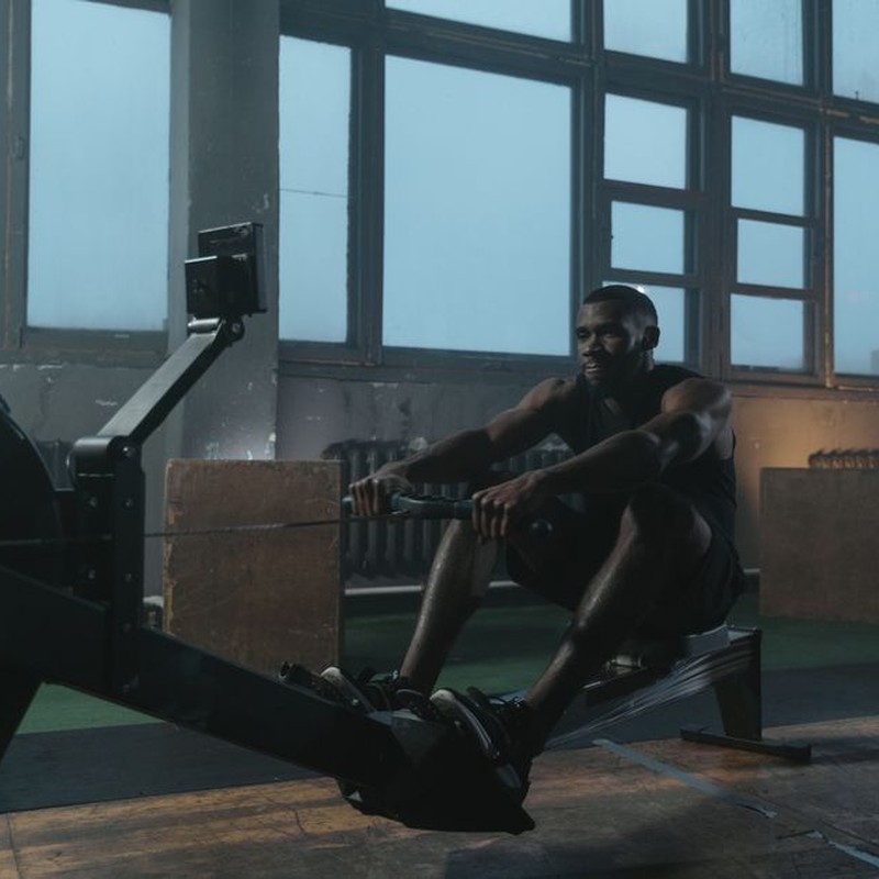 Focused athletic man working out on a rowing machine in a dimly lit gym setting.