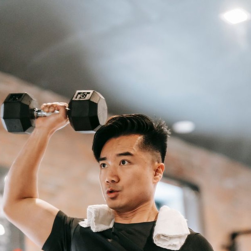 Powerful Asian male with towel standing with raised arm and lifting heavy dumbbell while pumping bicep in spacious gym on blurred background