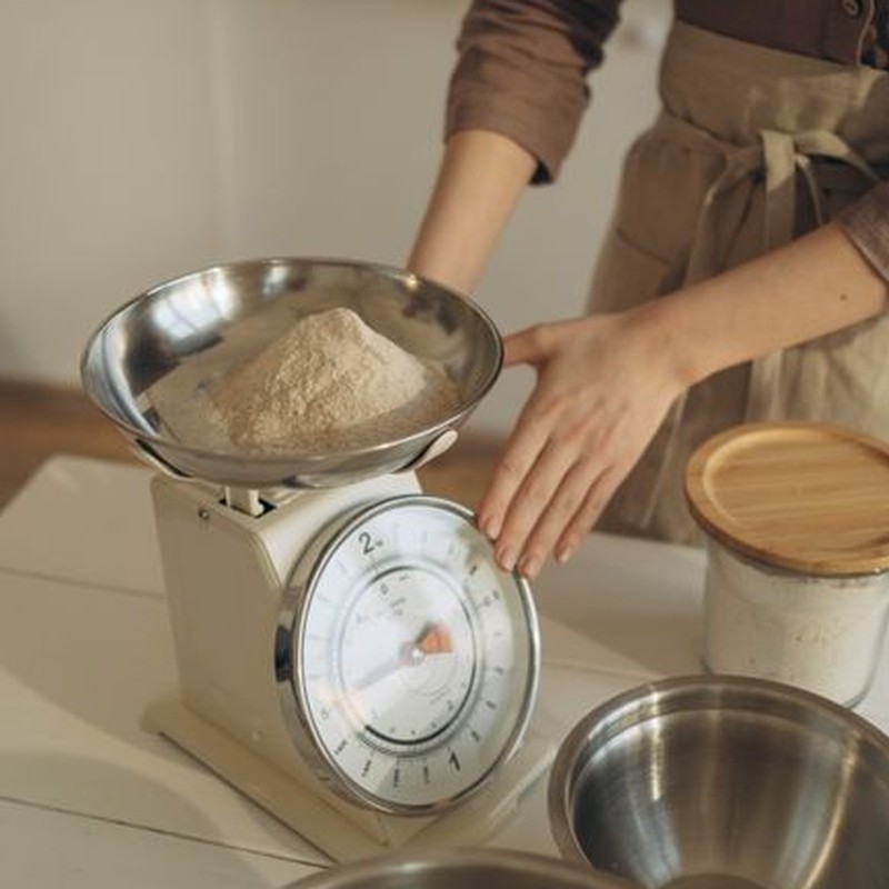 A woman weighing flour using a kitchen scale, preparing for baking in a cozy kitchen setting.