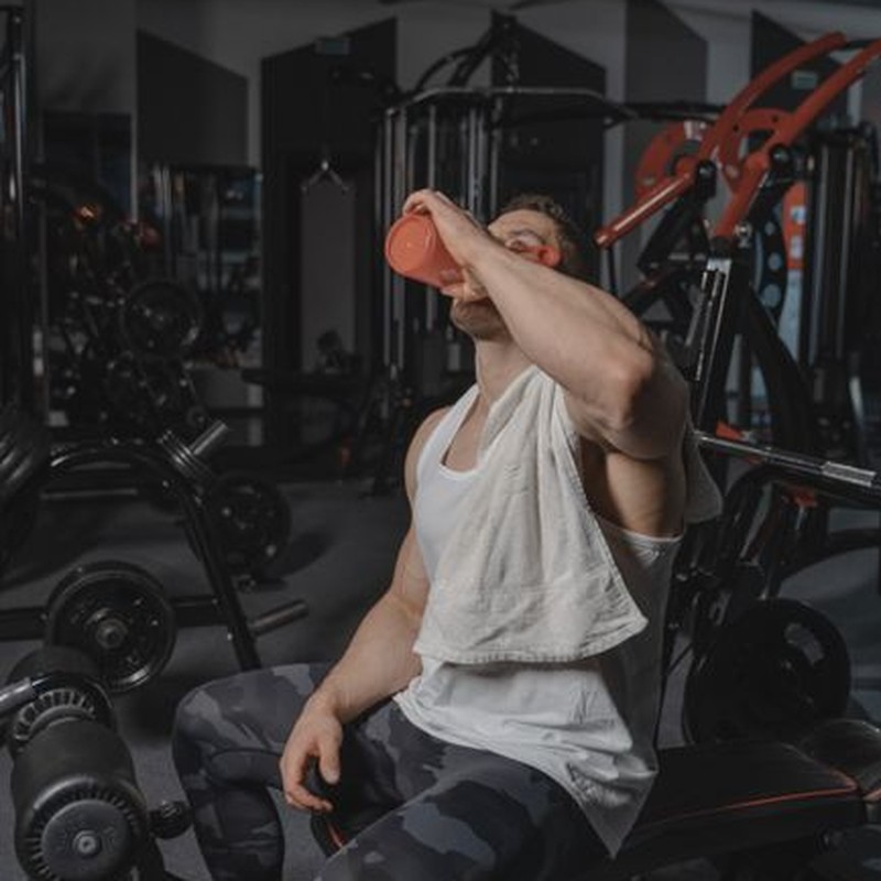 A muscular man with a towel rests and drinks water after an intense workout in a modern gym.