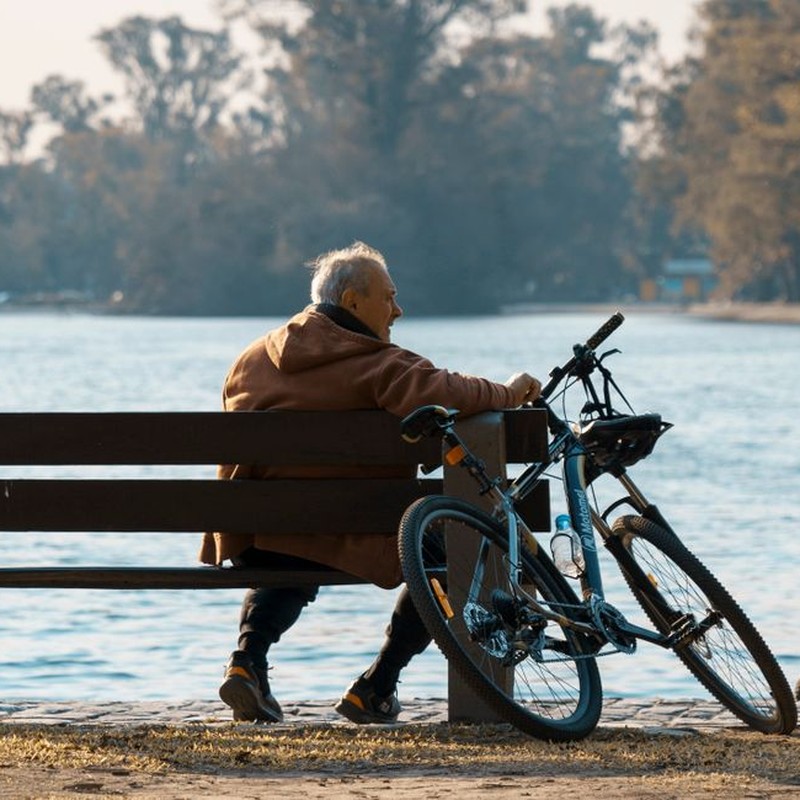 A senior man with gray hair sitting on a bench by the river, accompanied by his bicycle.