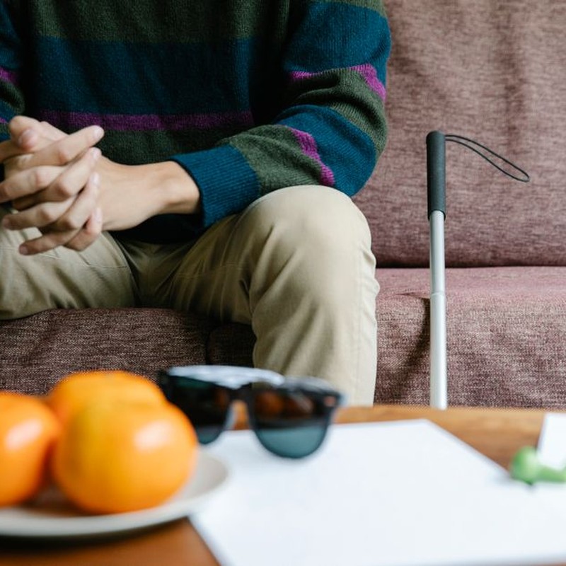Adult person with a walking stick sitting casually on a sofa indoors, with hands clasped.