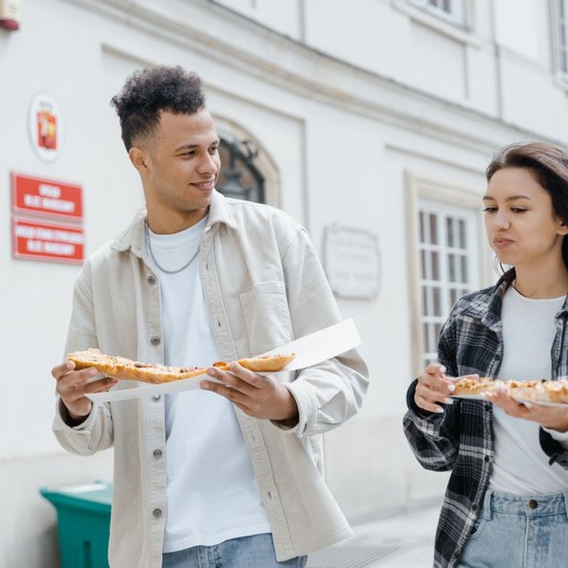 Two young adults enjoying pizza while walking outdoors in the city, creating a joyful street food moment.