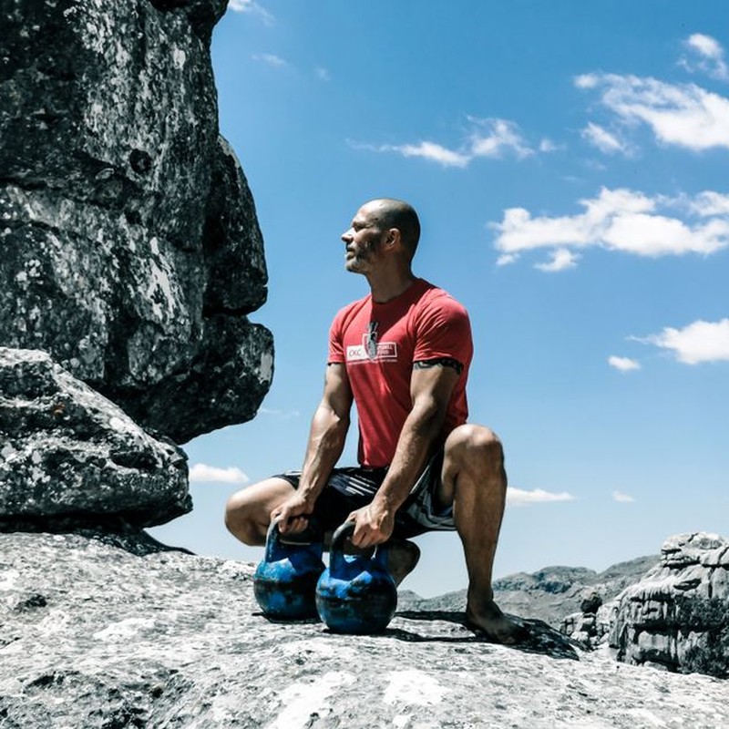 A muscular man exercises with kettlebells on rocky ground under a bright blue sky, emphasizing strength and fitness.