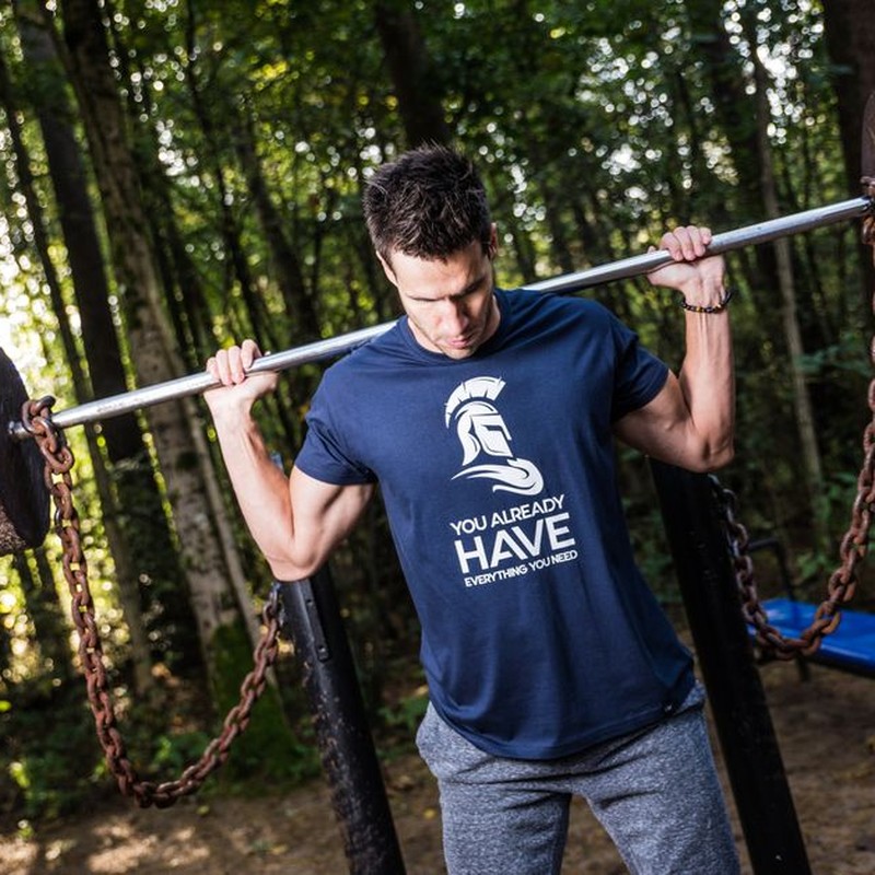 Man lifting barbell with chains in a wooded outdoor gym, showcasing strength training.