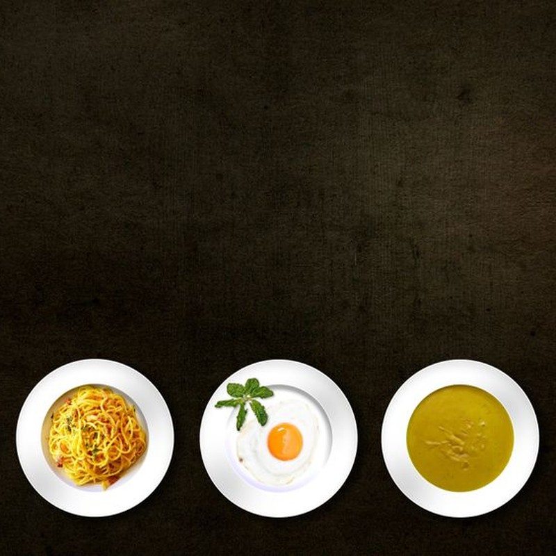 Flatlay of various dishes including pasta, egg, soup, and croissant on textured background.
