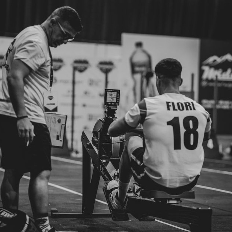 An athlete using a rowing machine indoors, coached in a gym setting.