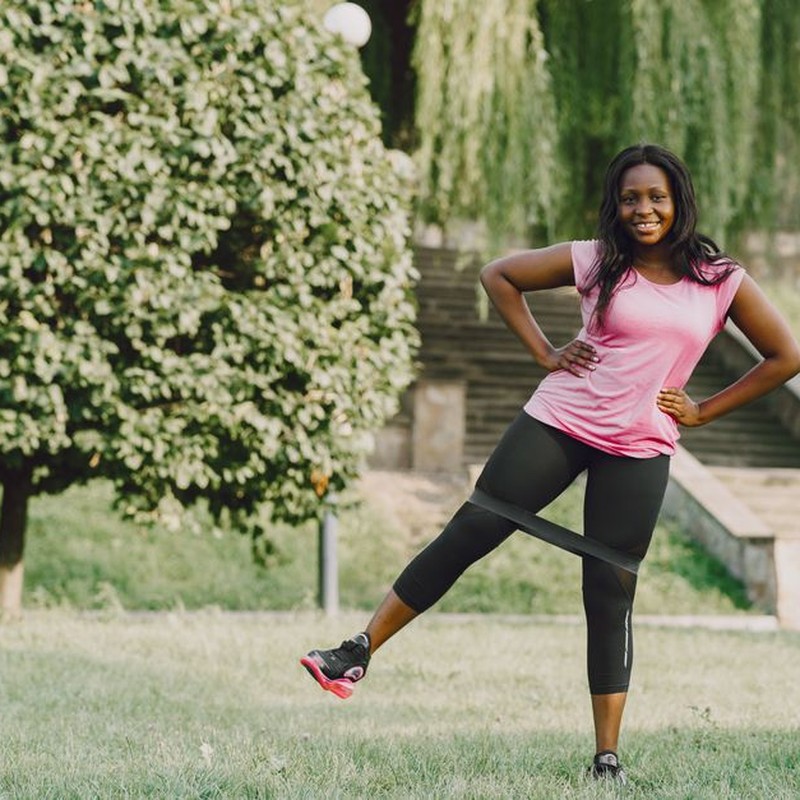 Smiling woman doing leg workouts with resistance band in a sunny park.
