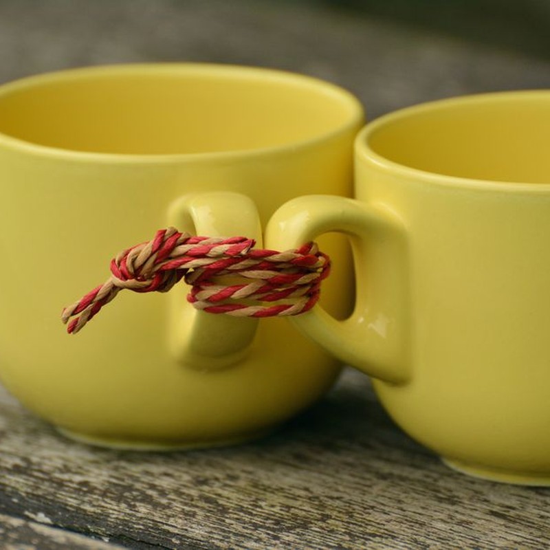 Two yellow ceramic mugs tied together with a red and yellow twine knot on rustic wooden surface.