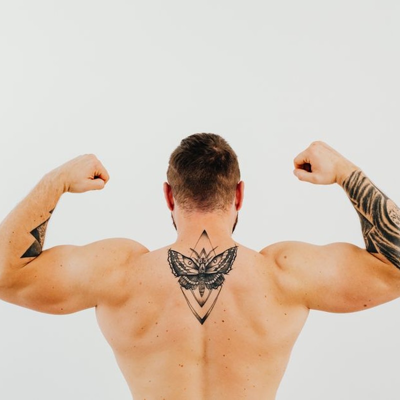 Shirtless man showcasing tattoos and biceps while flexing in a studio setting.