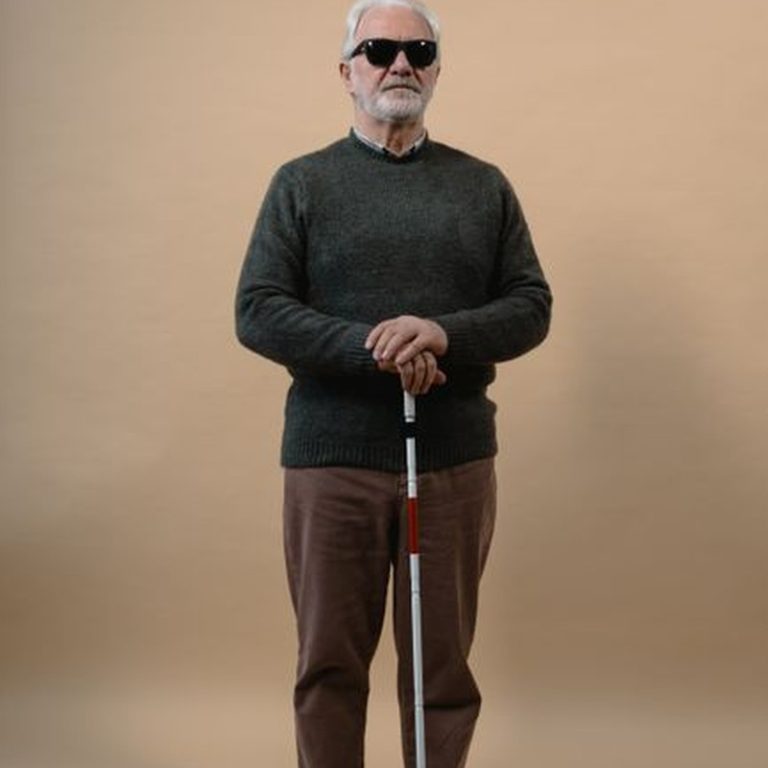 Senior man with visual impairment holding a cane, wearing sunglasses in a studio setting.