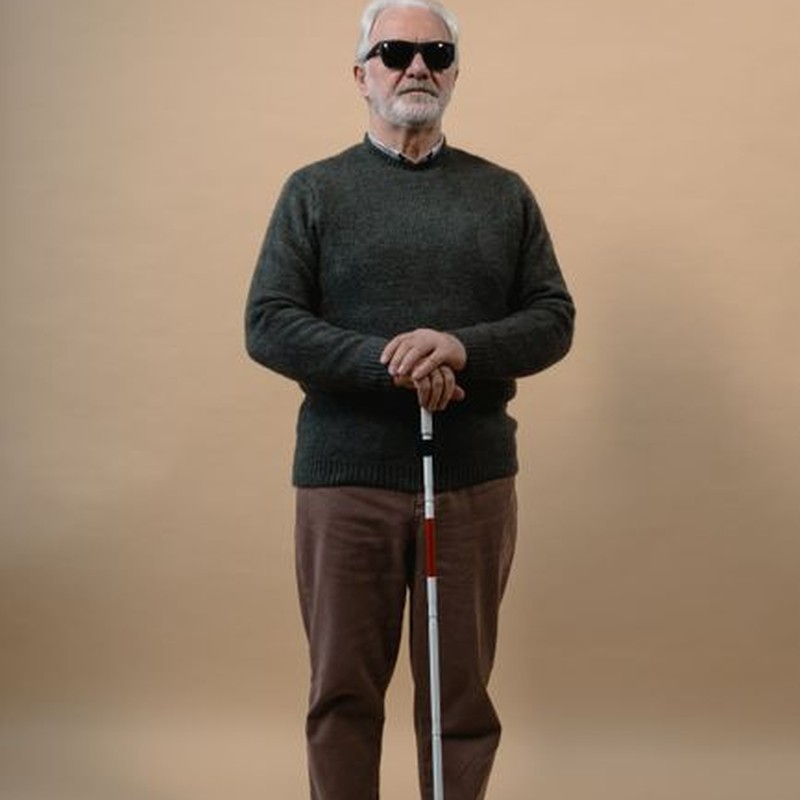 Senior man with visual impairment holding a cane, wearing sunglasses in a studio setting.