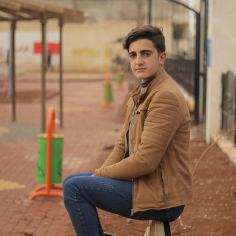 A young man sits on a bench outdoors in a casual setting, looking thoughtful.