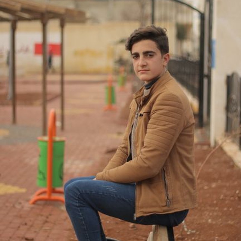 A young man sits on a bench outdoors in a casual setting, looking thoughtful.