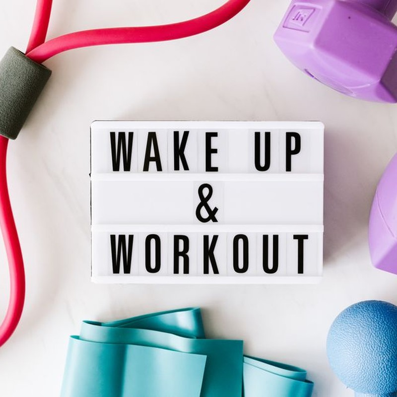 Top view closeup composition of wake up and workout inscription on light box surface among expanders and dumbbells and massage ball on white background