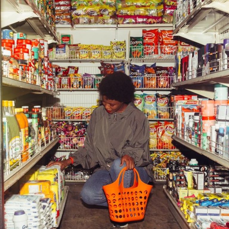 Full length of adult ethnic female buyer selecting goods while squatting with shopping bag among grocery shelves