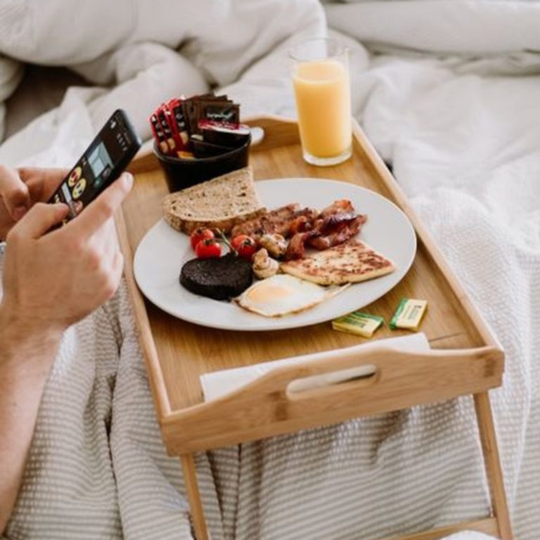 From above of crop unrecognizable person surfing internet on cellphone near tasty brunch on wooden tray at home