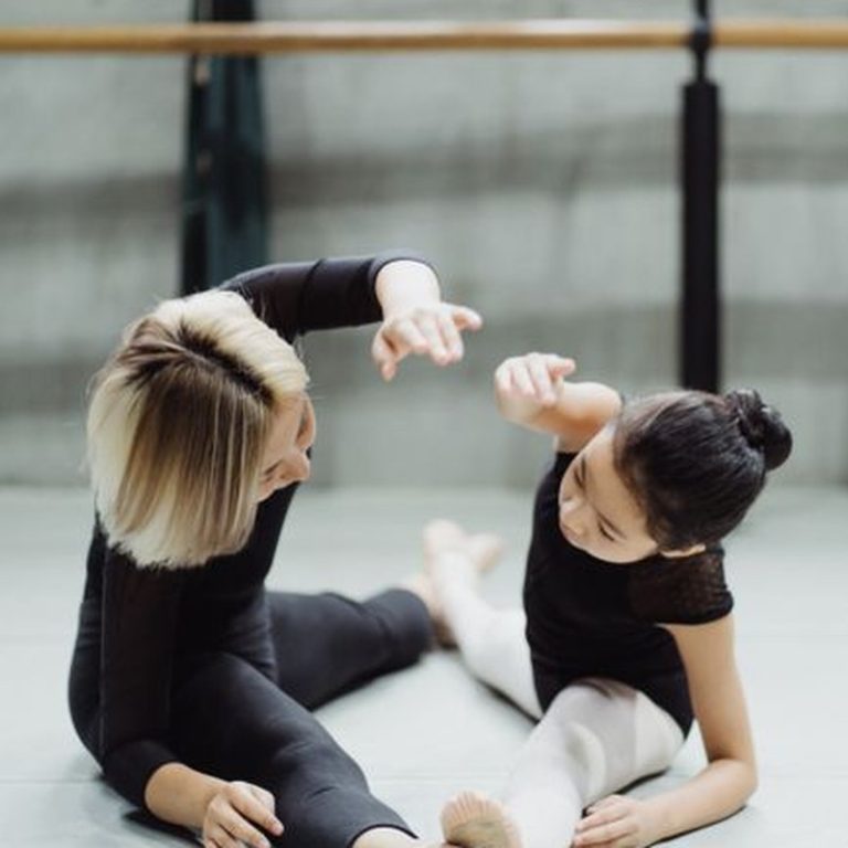 Side view of concentrated ballet teacher and focused girl stretching with raised hand together on floor in studio in daylight