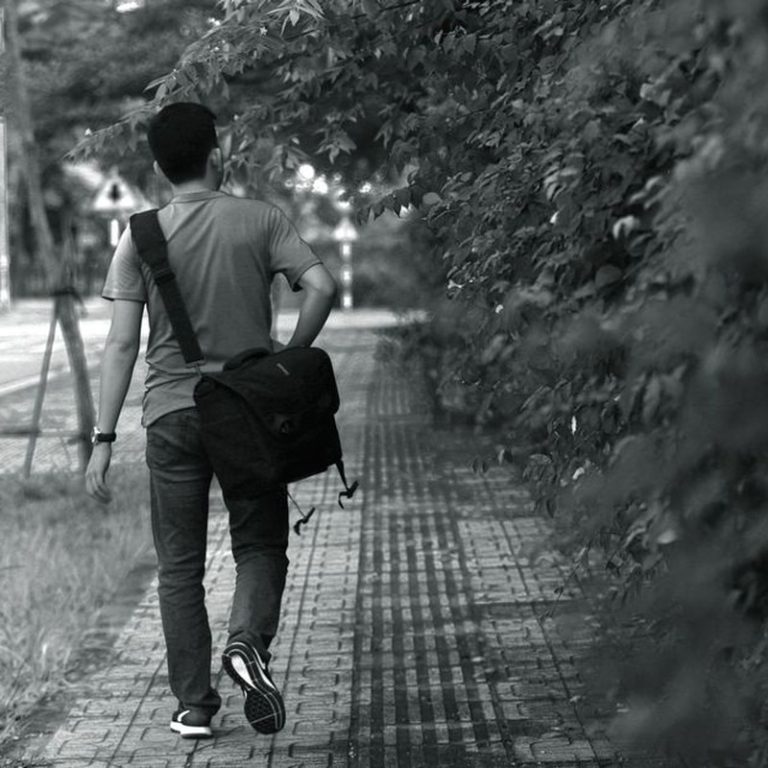 Black and white photo of man walking on sidewalk with messenger bag.