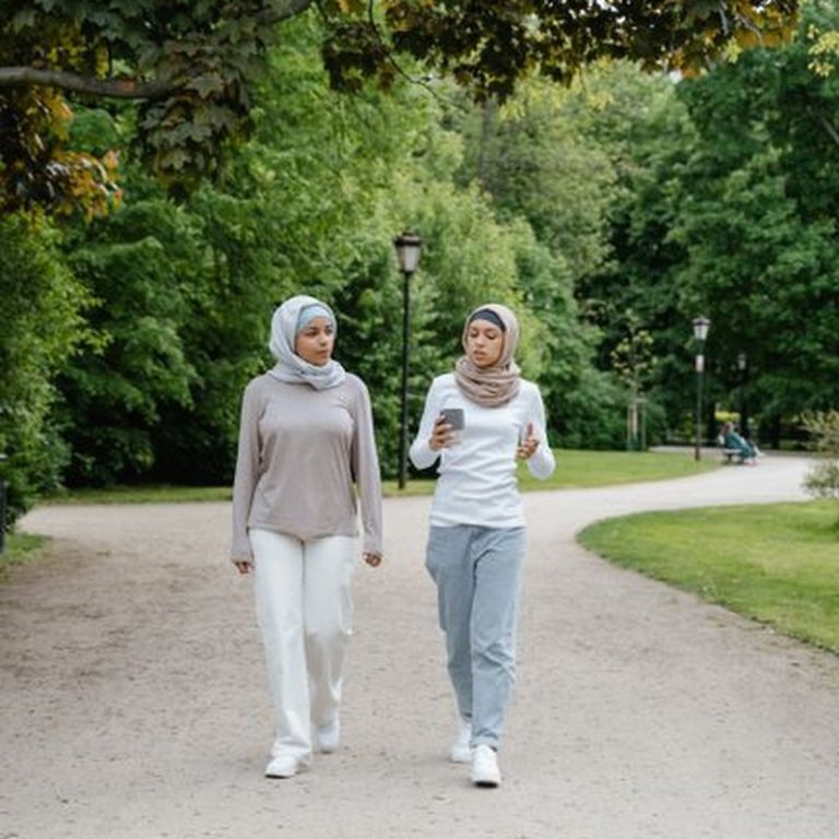 Two women wearing hijabs walk and converse in a lush green park setting.