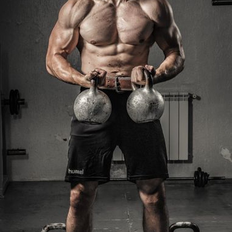 Shirtless man with muscular build lifting kettlebells indoors, showcasing strength and fitness.