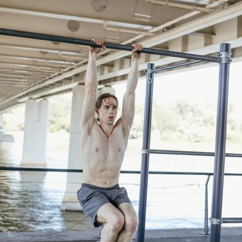 Shirtless man exercising on a pull-up bar in an outdoor gym under a bridge.