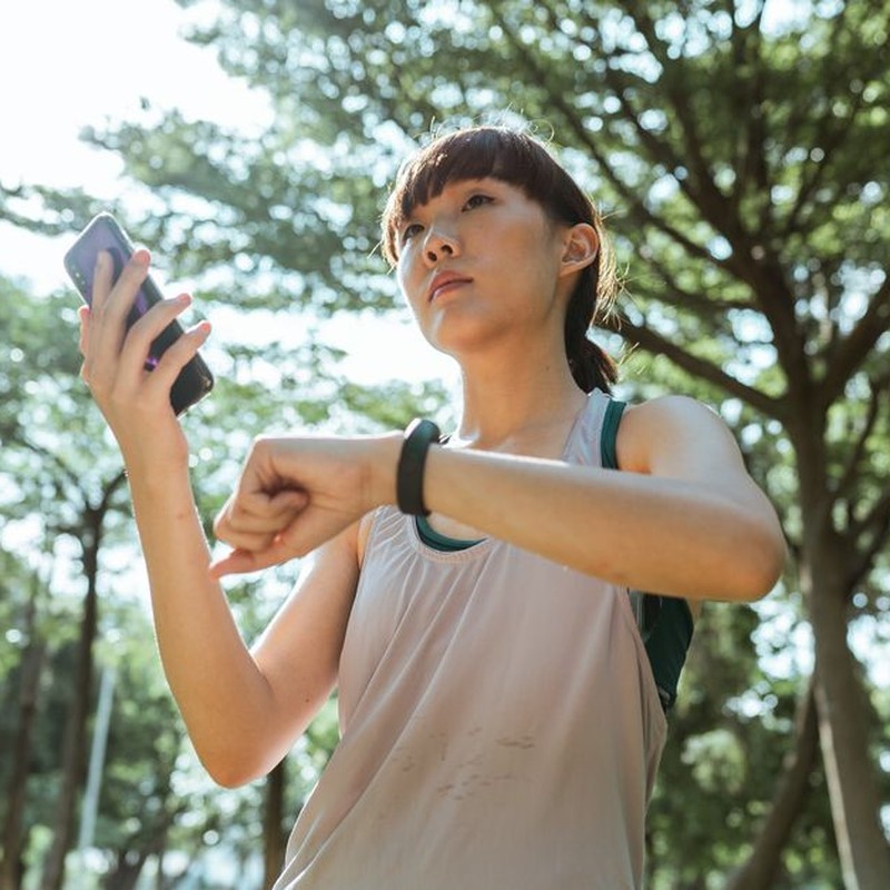From below ethnic Asian female wearing sport clothes standing in park and connecting smartphone and smart watch during break in sunny day