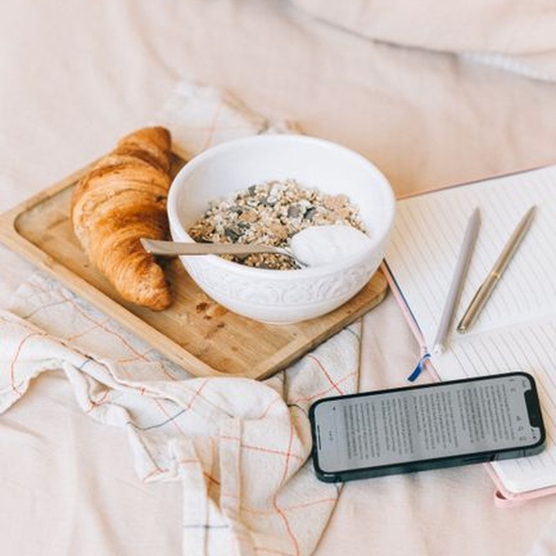 A tray of croissant and yogurt with granola on a bed, accompanied by a notebook and smartphone.