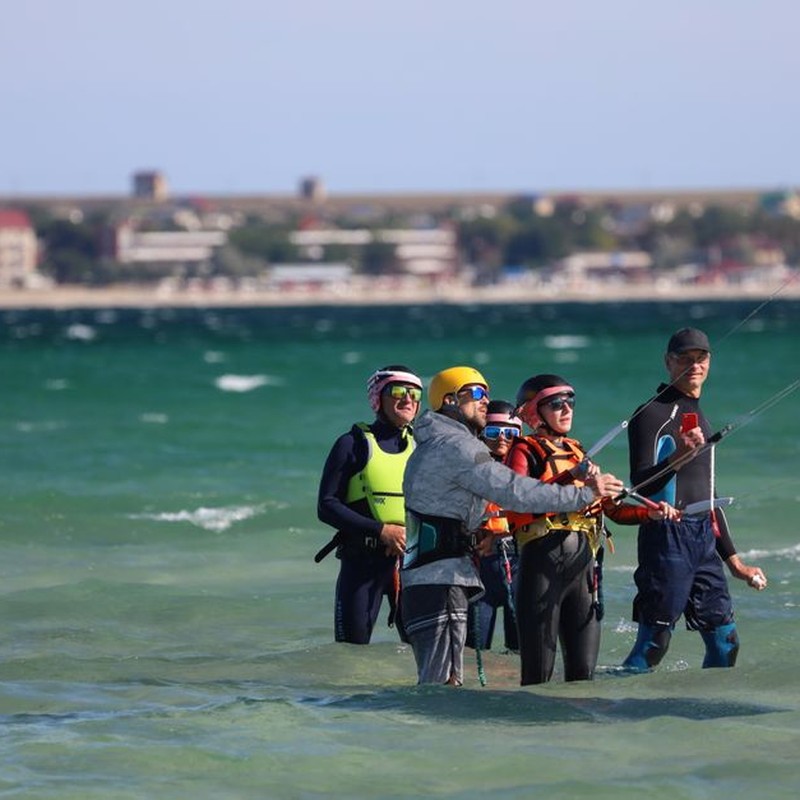 A group of people receiving a kiteboarding lesson in shallow sea waters, equipped with helmets and gear.