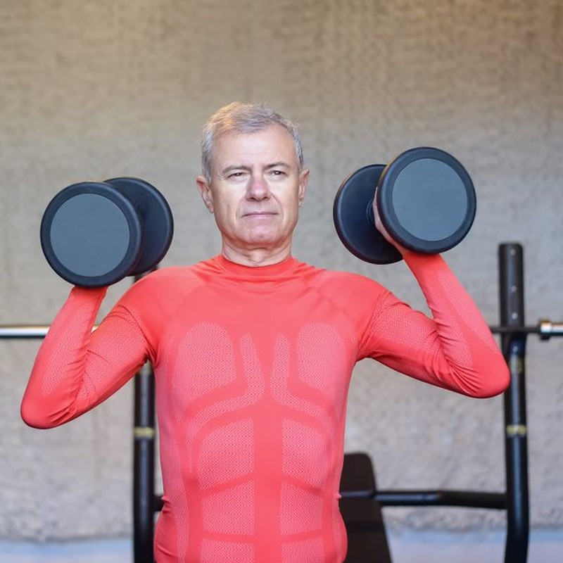 Senior man in vibrant red outfit weightlifting indoors, promoting fitness and healthy lifestyle.