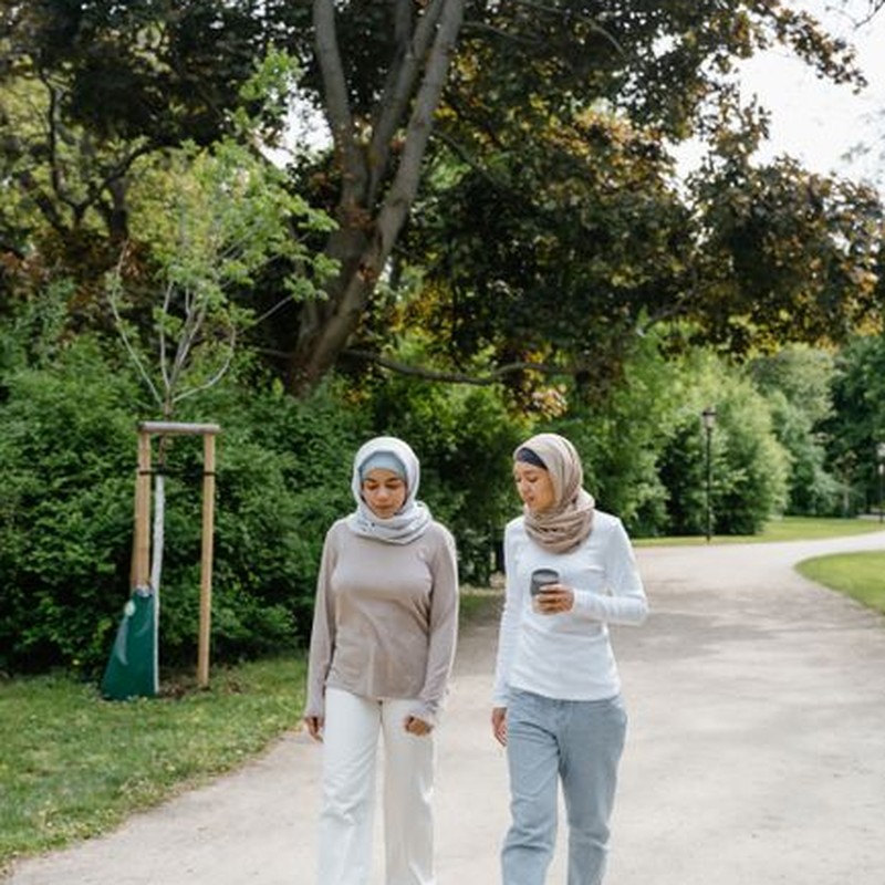 Two women wearing hijabs walking on a pathway through a green park in daylight.