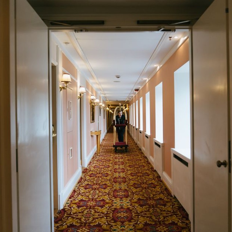 A bellboy pushes a luggage cart down a luxurious hotel corridor with elegant lighting and patterned carpet.