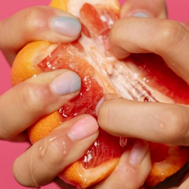 Vibrant grapefruit juice being squeezed by hands with pastel nails on pink background.