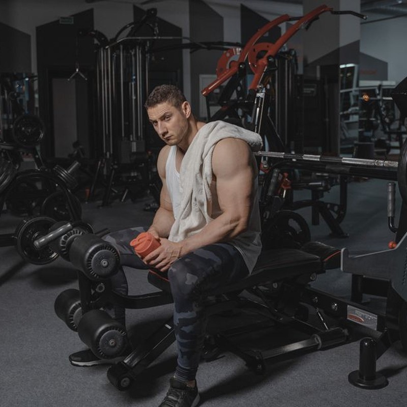 Man seated on bench in gym holding protein shaker, surrounded by equipment.