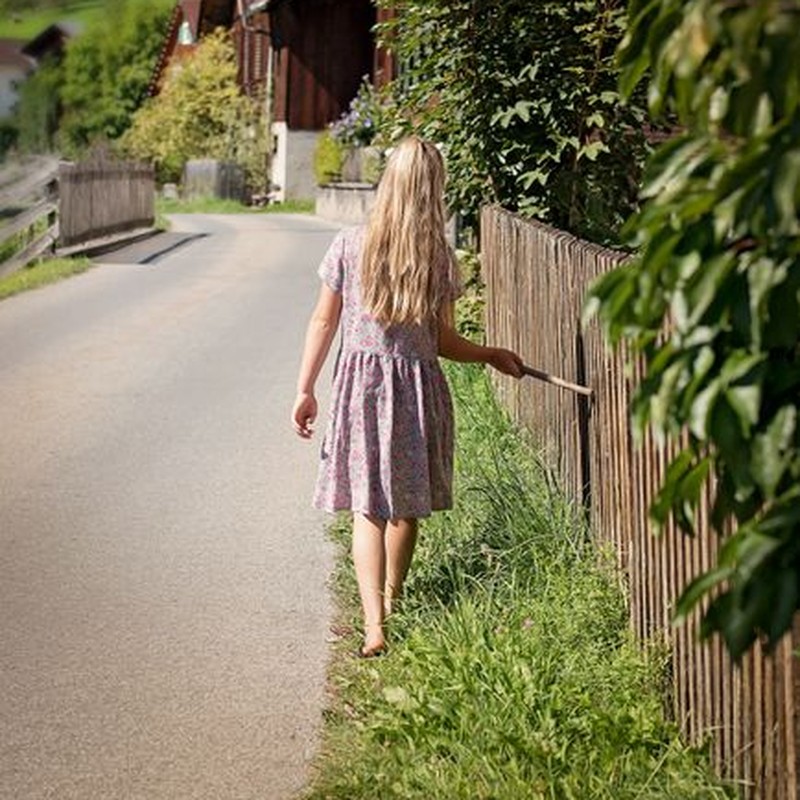 A young girl walks barefoot on a rural road, touching a fence on a sunny day.