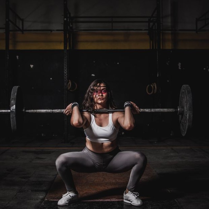 A strong female weightlifter squats with a barbell in a dimly lit gym, showcasing power and fitness.