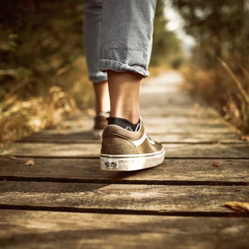 Person wearing sneakers walking on a wooden path in a sunlit park.