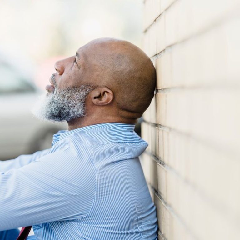 Side view of a thoughtful man with a beard leaning against an outdoor brick wall.