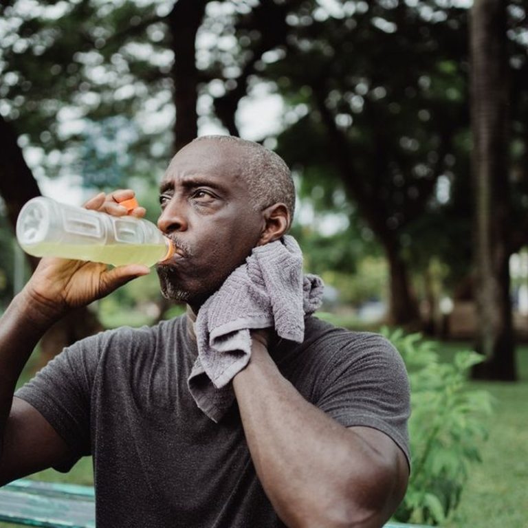 Adult man drinking sports beverage and wiping sweat in a park.