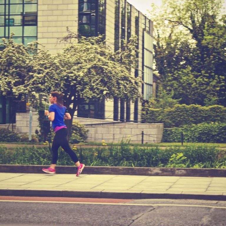 A woman running along a city street during the day surrounded by nature.