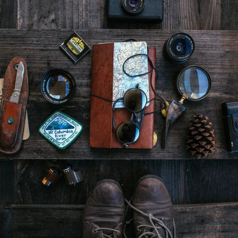 Overhead of vintage photo camera near leather notebook with map and knife near camera roll and lenses near sunglasses placed on table near boots